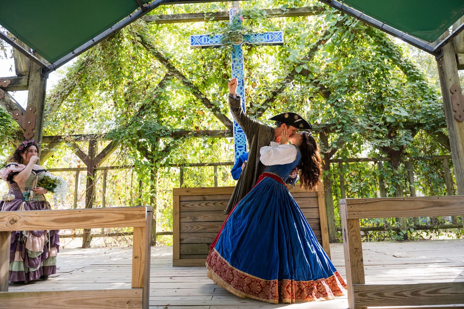 A group of dancers in historical costumes perform traditional dances under a wooden pavilion in a park setting.