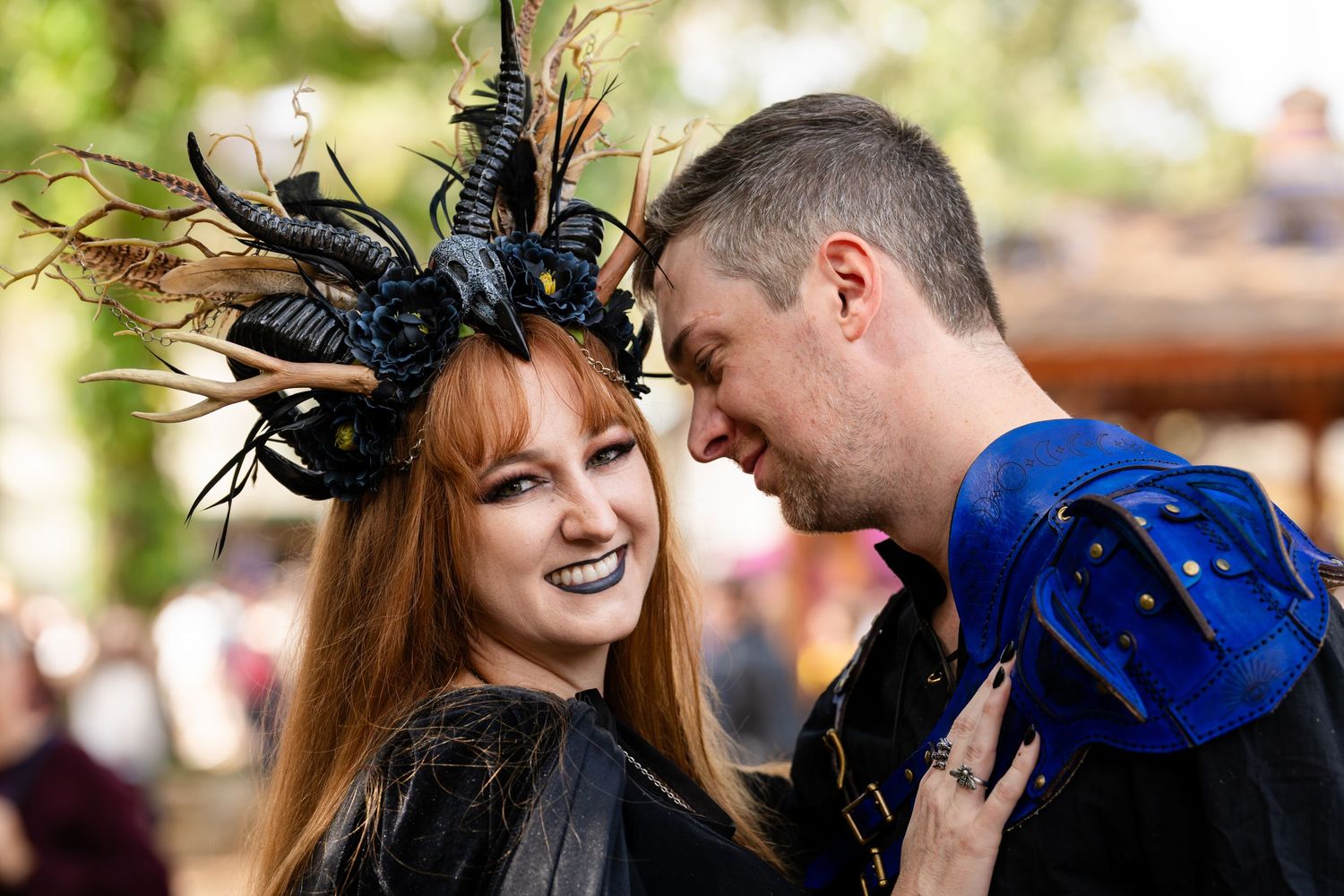 A couple in Renaissance festival attire share a moment, with one wearing an elaborate feathered headpiece and black dress.