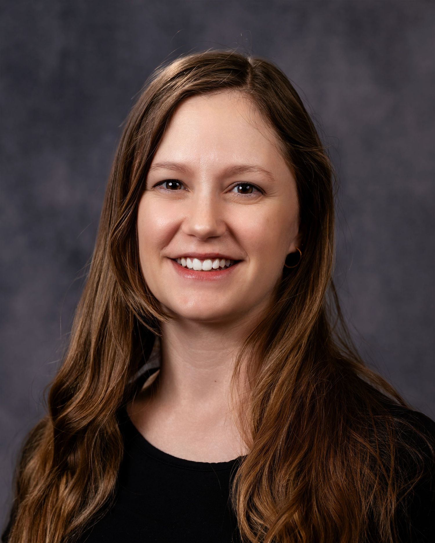 Professional headshot portrait of someone with long brown hair and a warm smile against a dark gray background.
