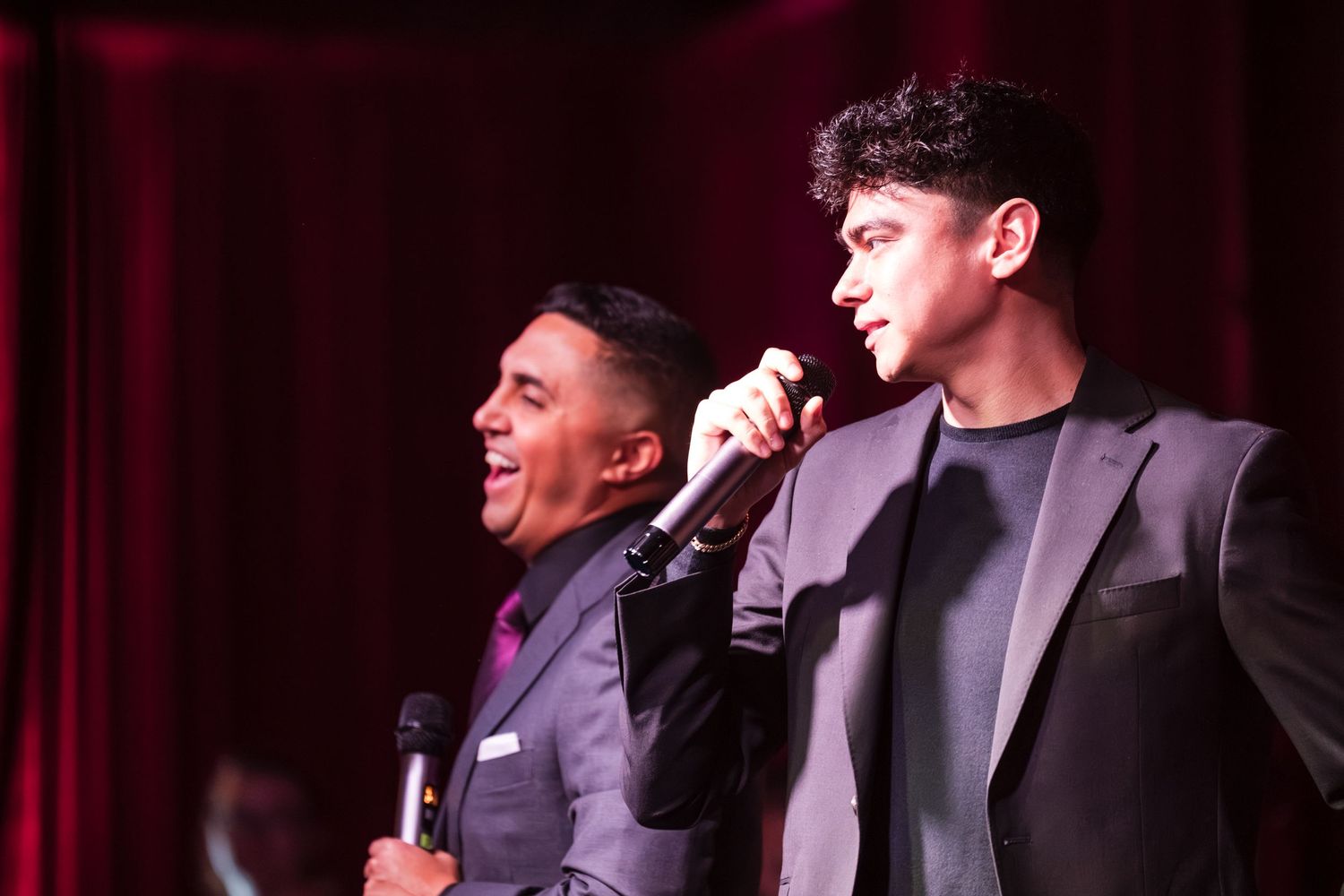 Performers in dark attire stand on stage with microphones against a deep red curtain backdrop during a show.