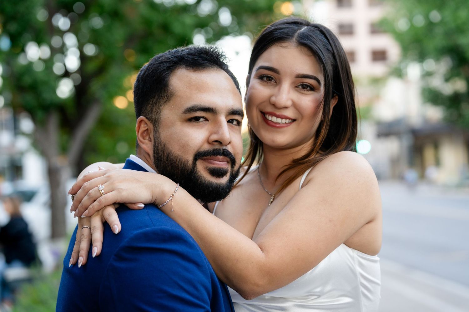 Happy couple embraces outdoors with greenery and urban setting in background during engagement photoshoot.
