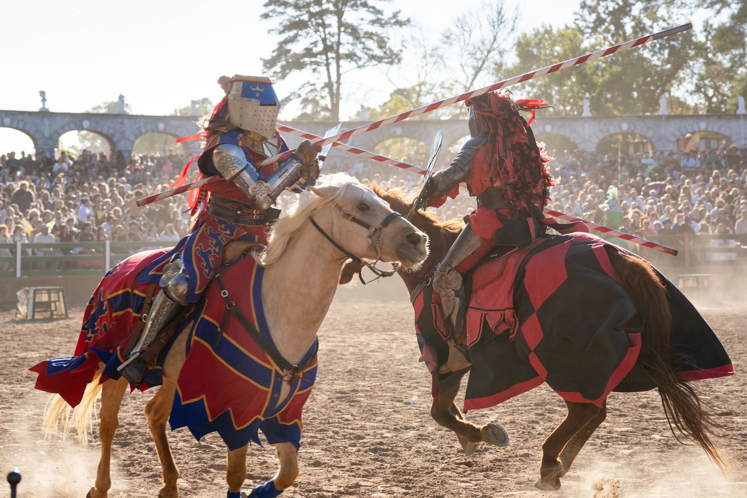 Two knights in medieval armor joust on horseback at a renaissance festival as a crowd watches through the dust.