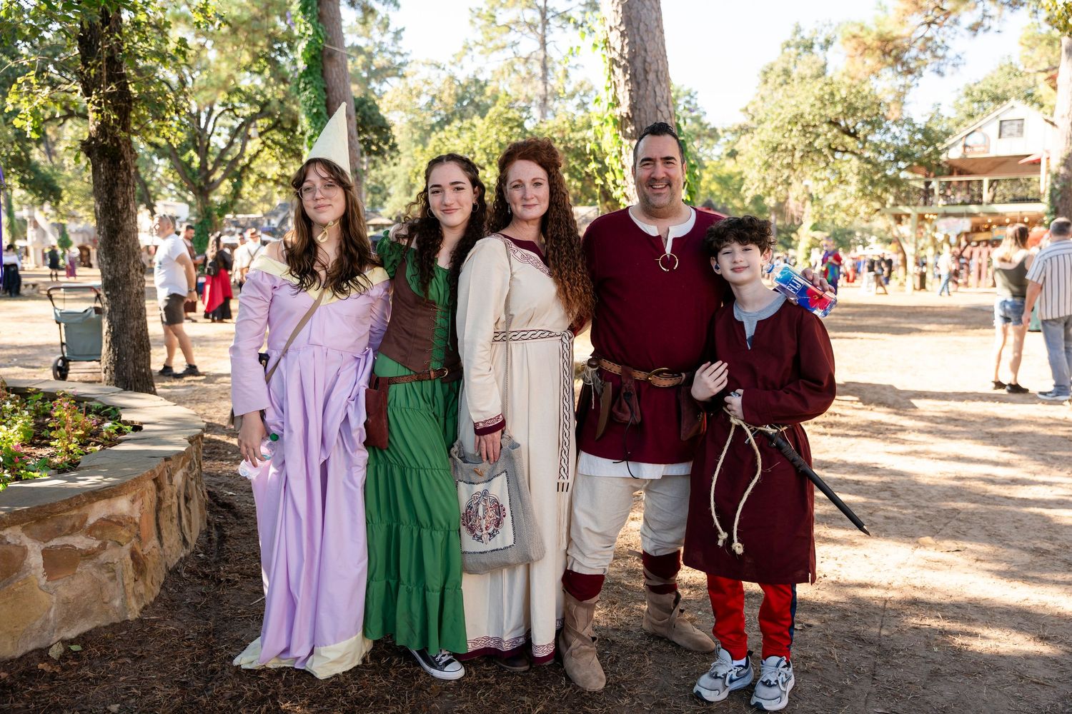 Group in medieval renaissance fair costumes standing together at an outdoor festival in a wooded area.