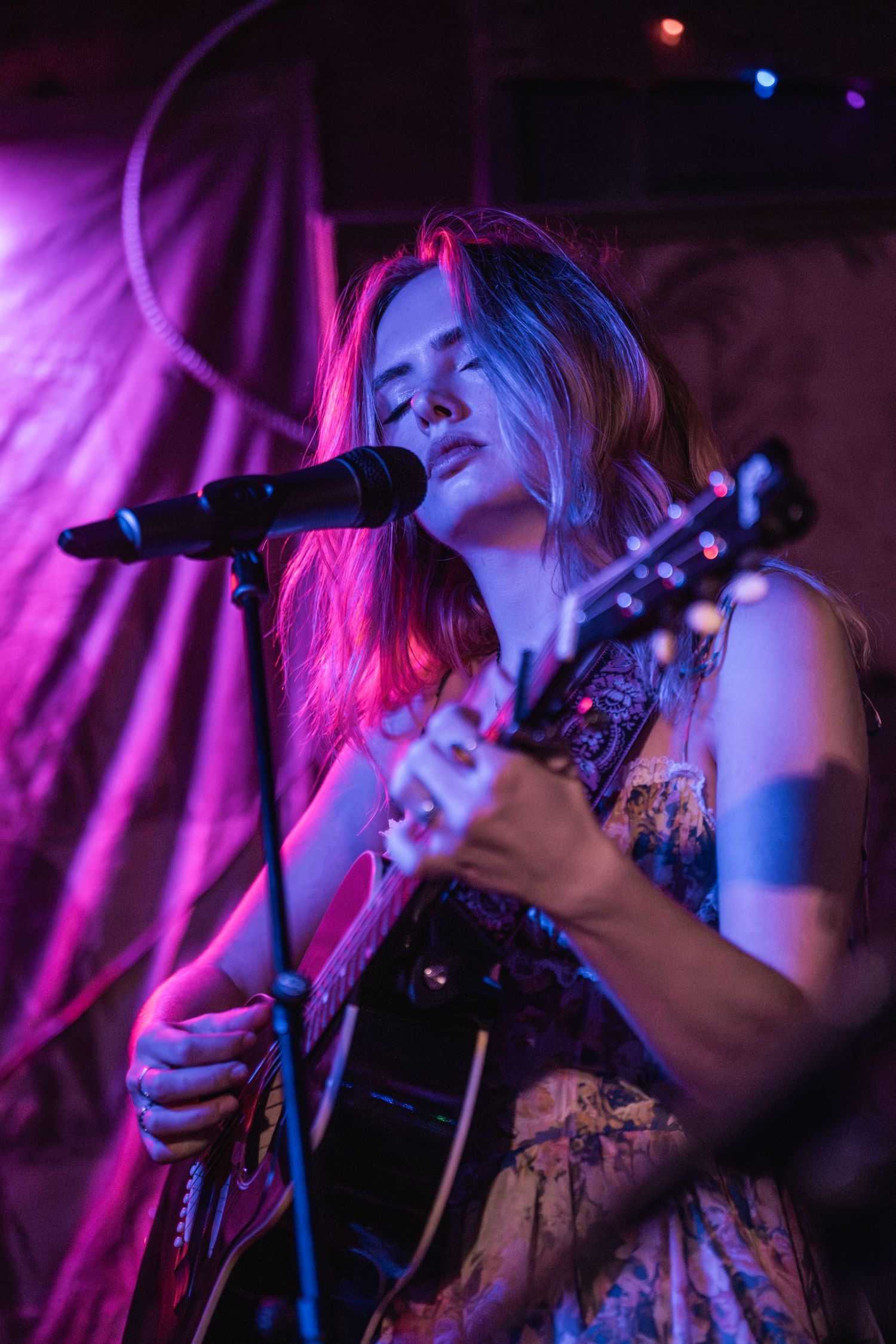 A musician performs on stage with an acoustic guitar under dramatic purple and pink lighting at a live music venue.