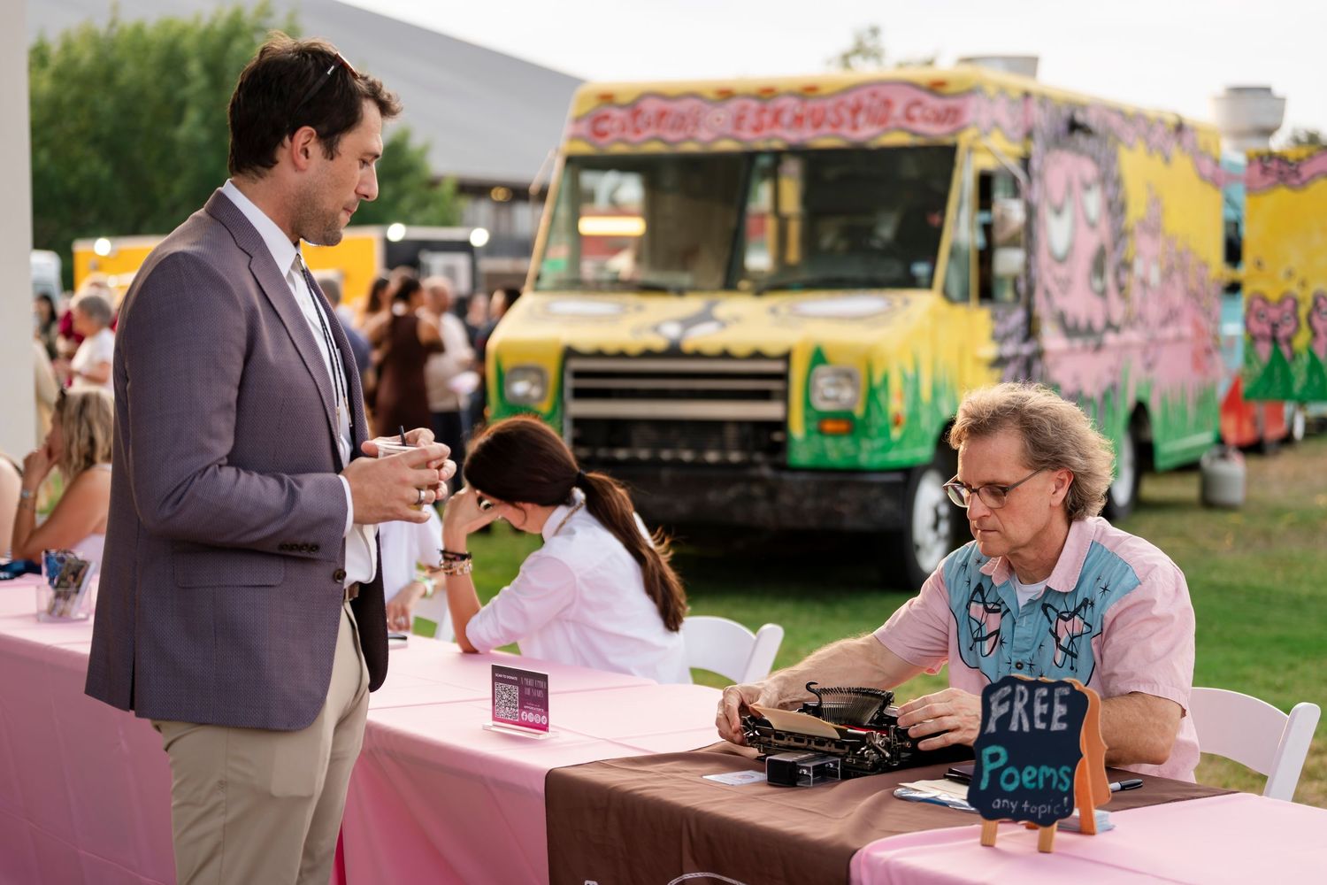 People interact at an outdoor book signing event with colorful food trucks in the background.