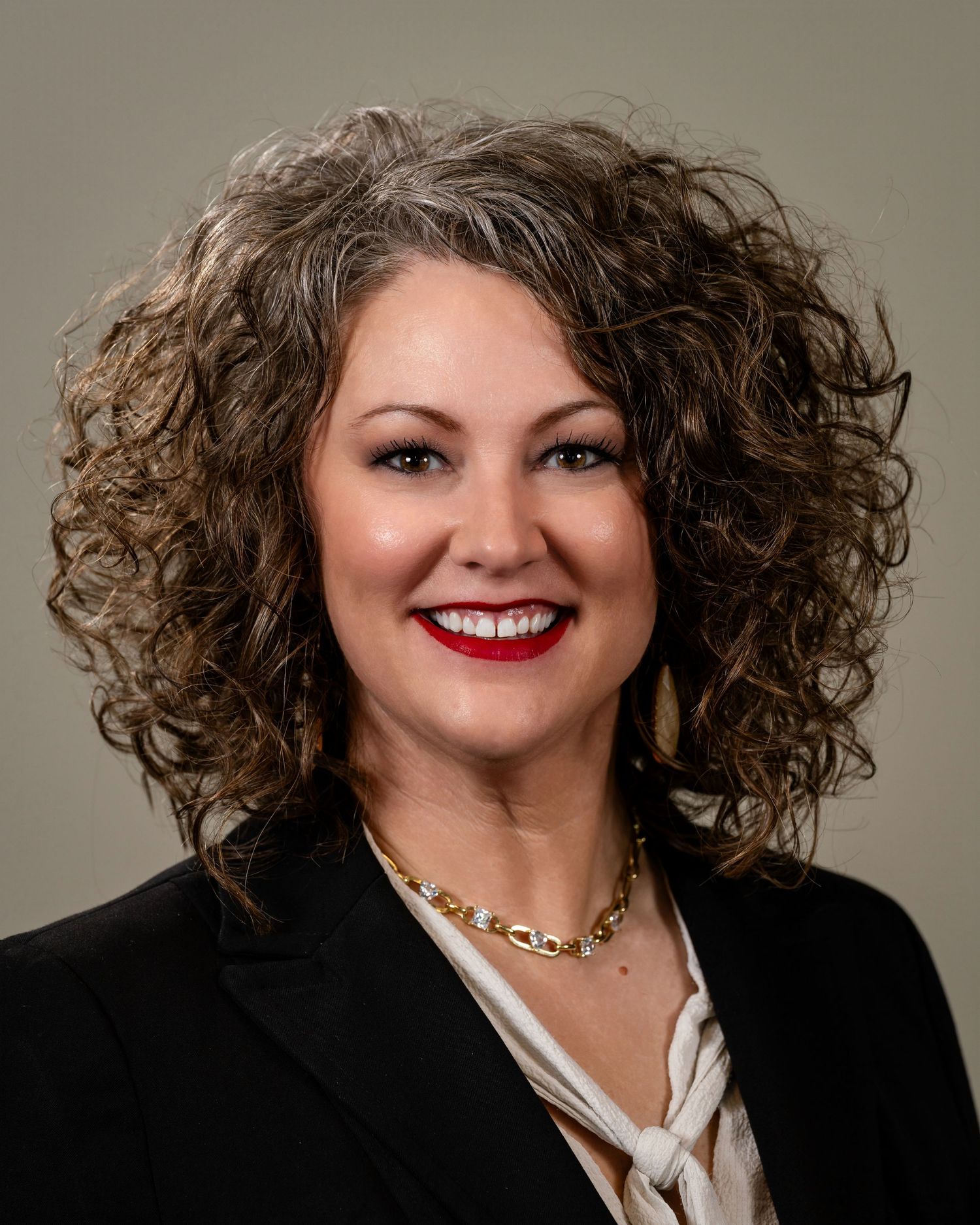 Professional headshot of a woman with curly brown and gray hair, red lips, gold necklace, and black blazer.