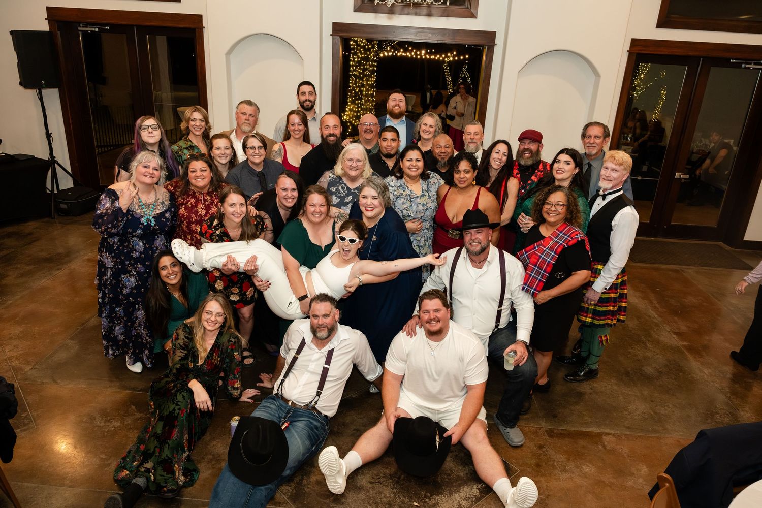 Large wedding group photo with bride and groom surrounded by smiling guests in formal attire at an elegant indoor venue.