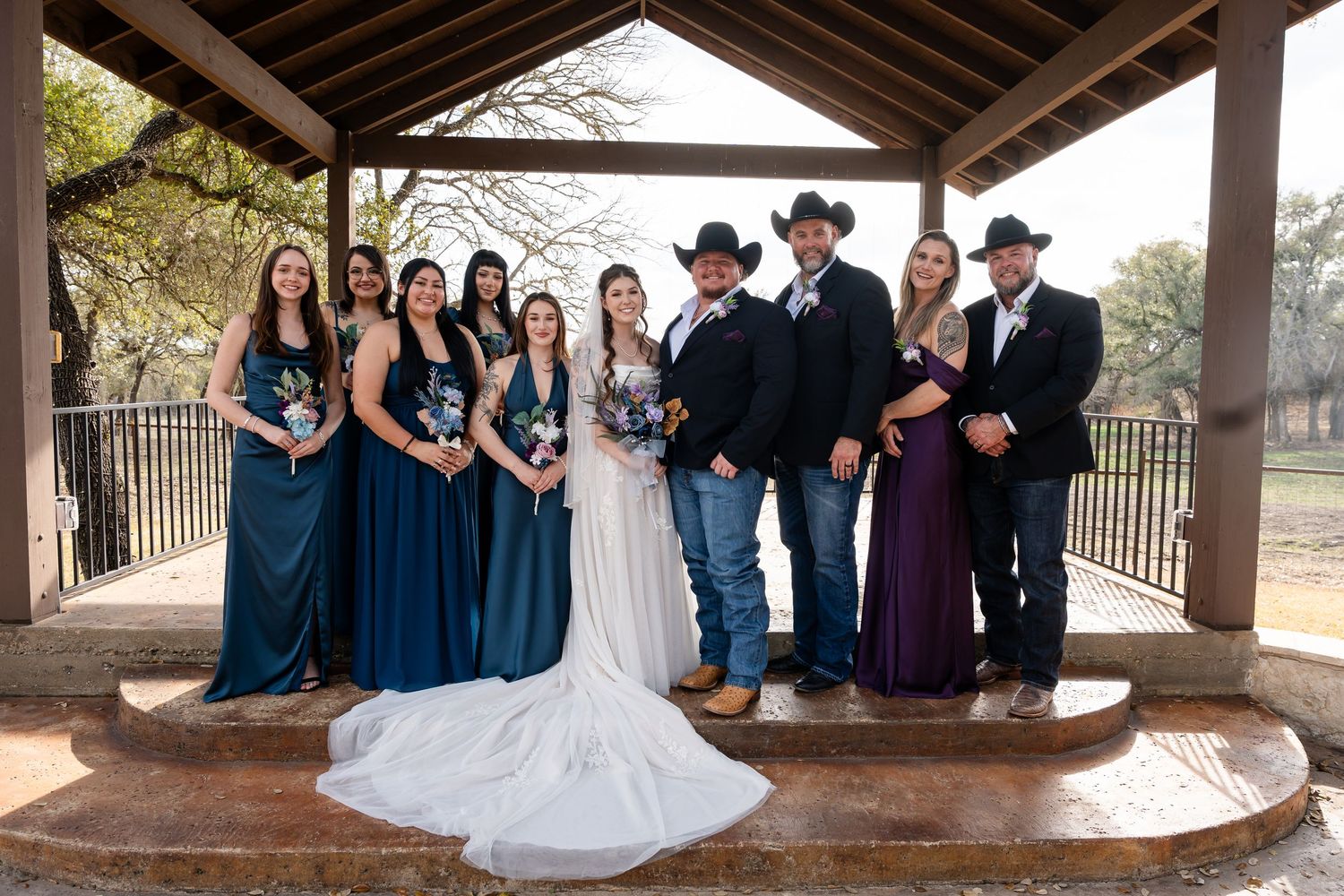 Wedding party poses under rustic pavilion, bridesmaids in teal gowns, groomsmen in cowboy hats, bride in white gown.