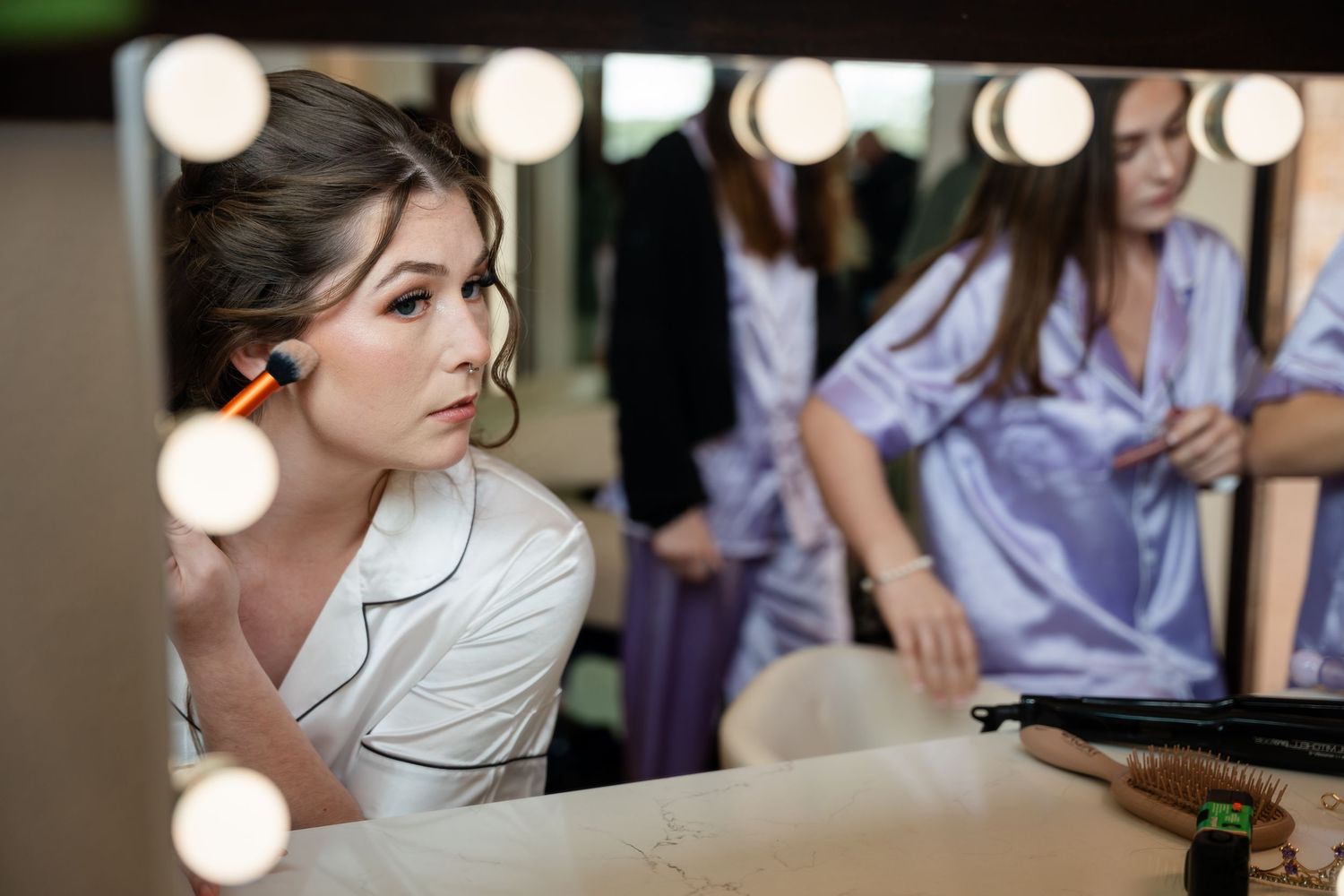 Bride in white satin robe getting ready at vanity mirror with bridesmaids in purple robes behind her.