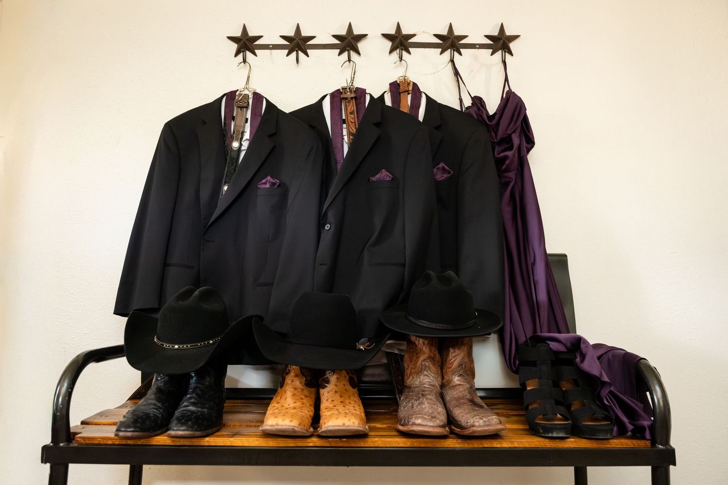 Three black suits with colorful shirts hang on a star rack above cowboy boots and hats on a wooden bench.