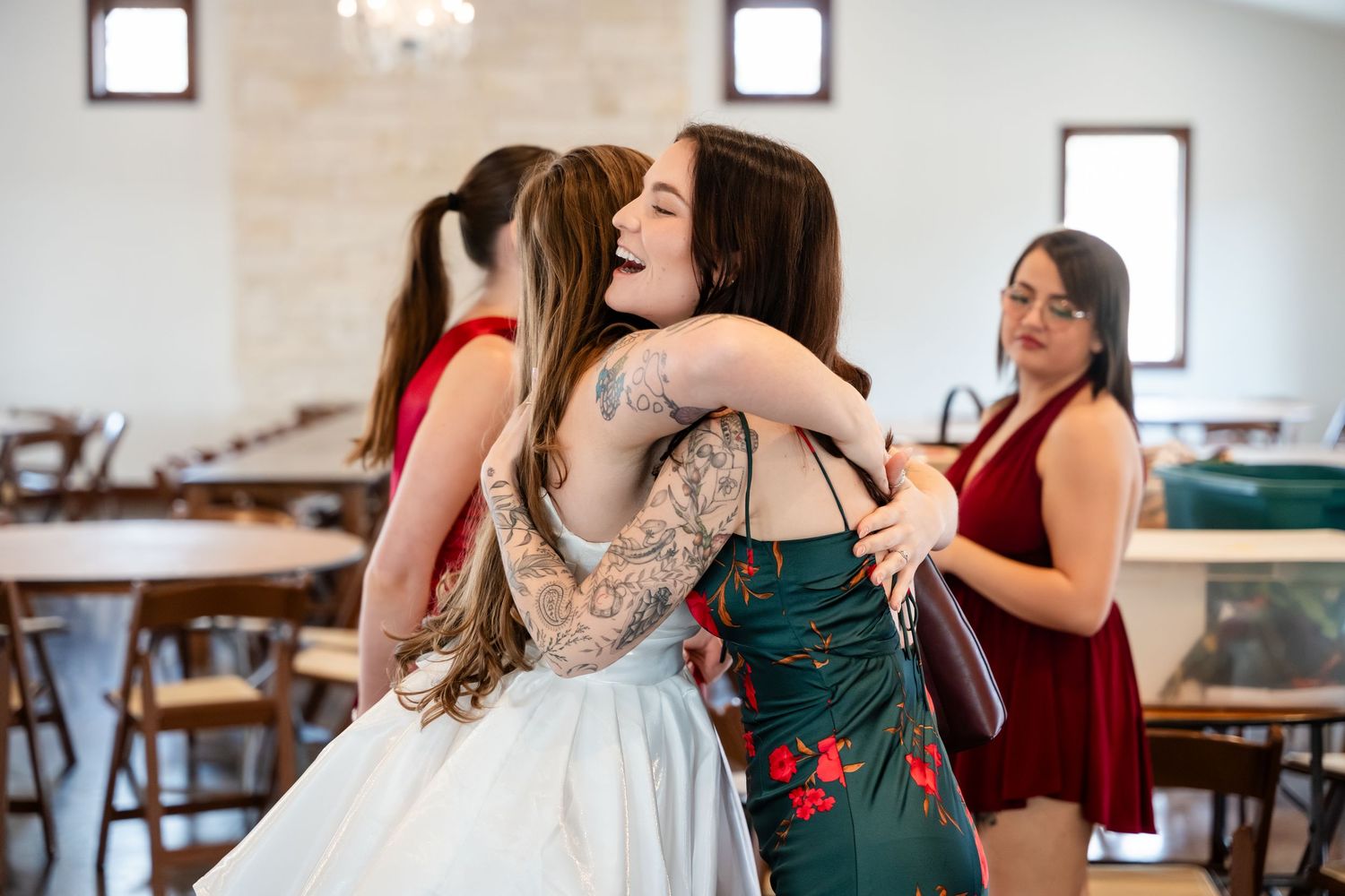 Two women share a warm hug at a wedding venue, one in a white gown and the other in a green floral dress.