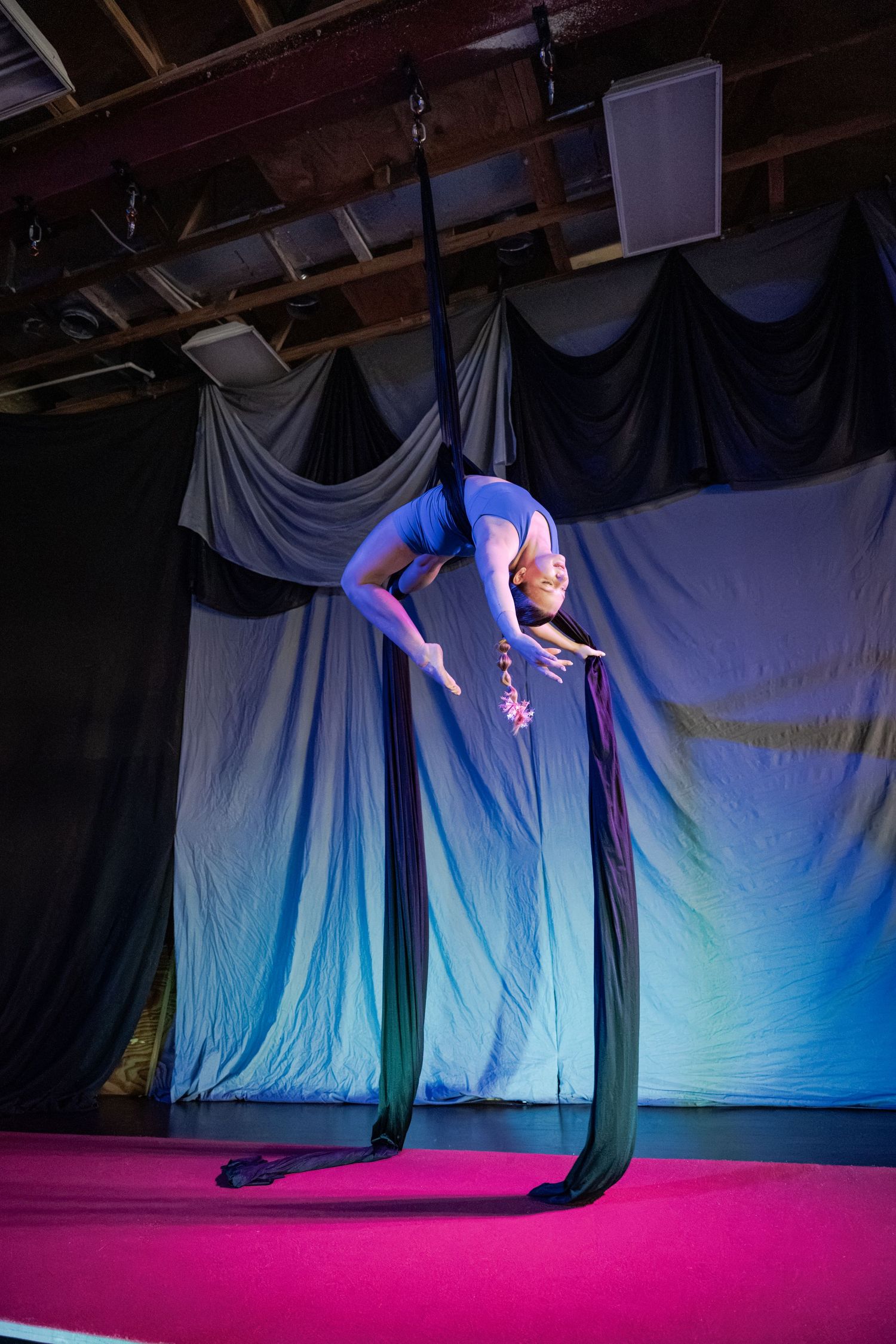 Aerial silk performer in blue leotard gracefully bending backward on dark fabric silks on a pink stage.