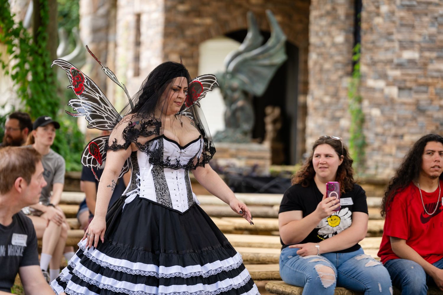 A performer in a black and white fairy costume with wings dances at a Renaissance faire for an audience.