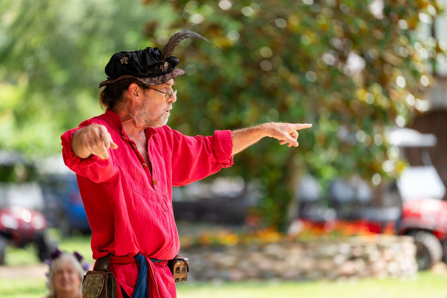 Man dressed as a pirate in red shirt and black hat pointing finger outdoors at a Renaissance fair.