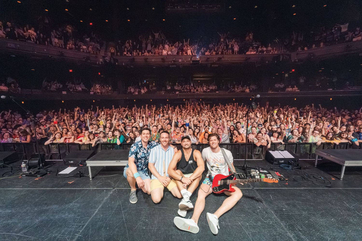 Four performers pose on stage with a packed concert hall audience behind them under warm venue lighting.