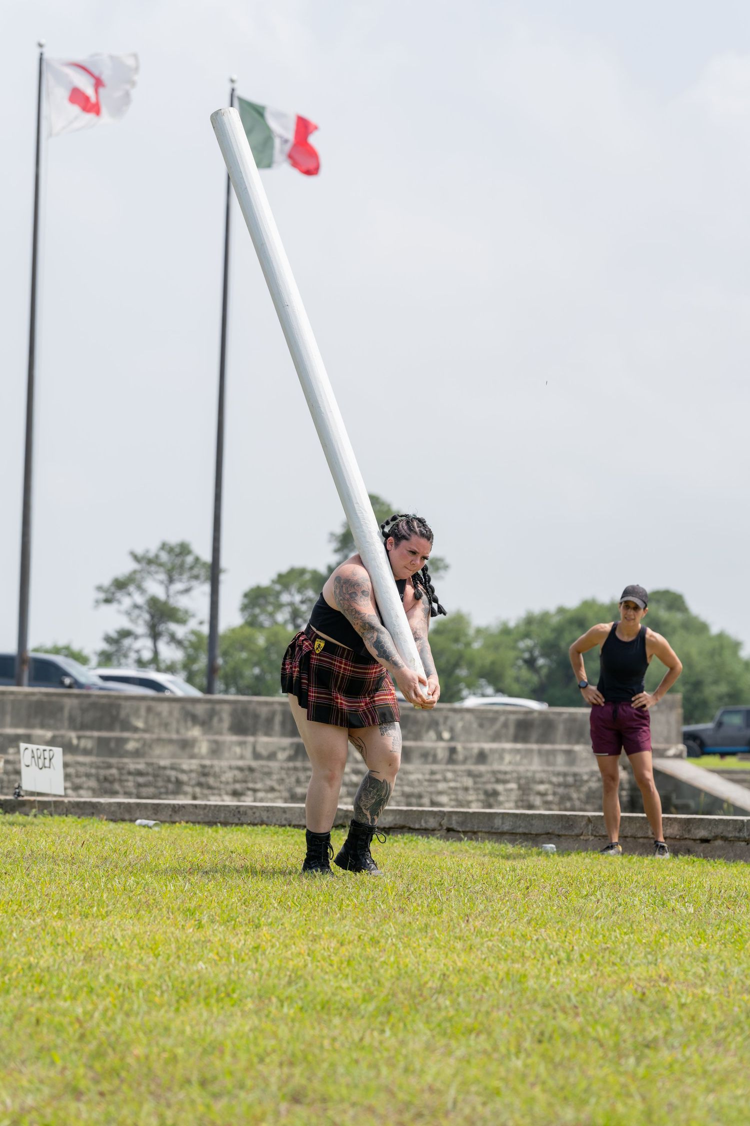 athlete in tartan kilt tossing caber at Scottish Highland Games competition on green field with flags in background.
