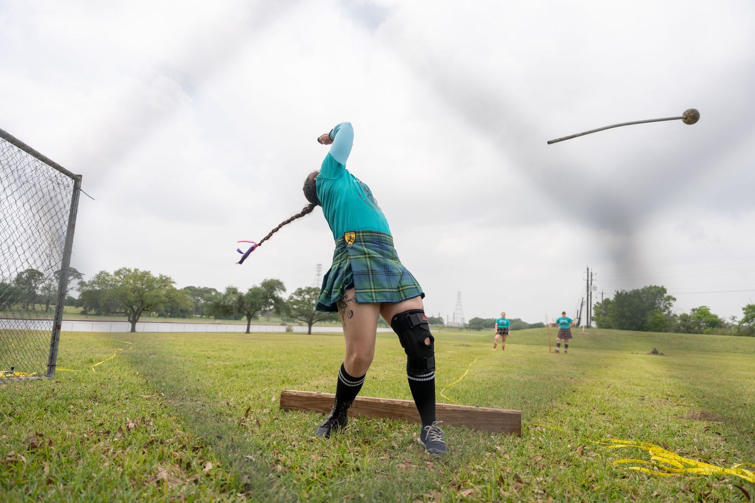 Athlete in teal and kilt throws hammer at Highland Games competition on grass field.