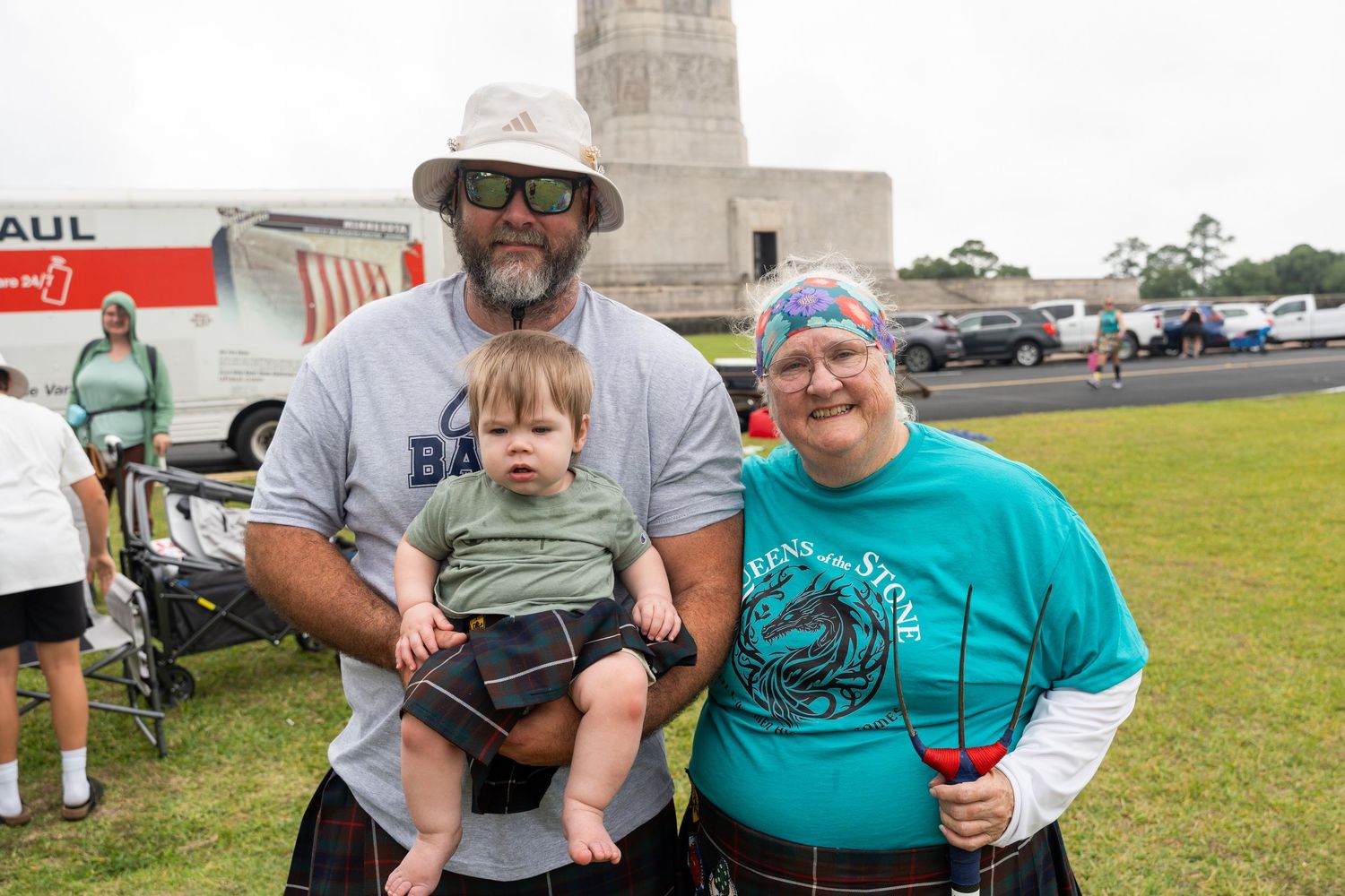 Family of three wearing kilts at outdoor Scottish Highland Games festival, with a U-Haul truck in background.