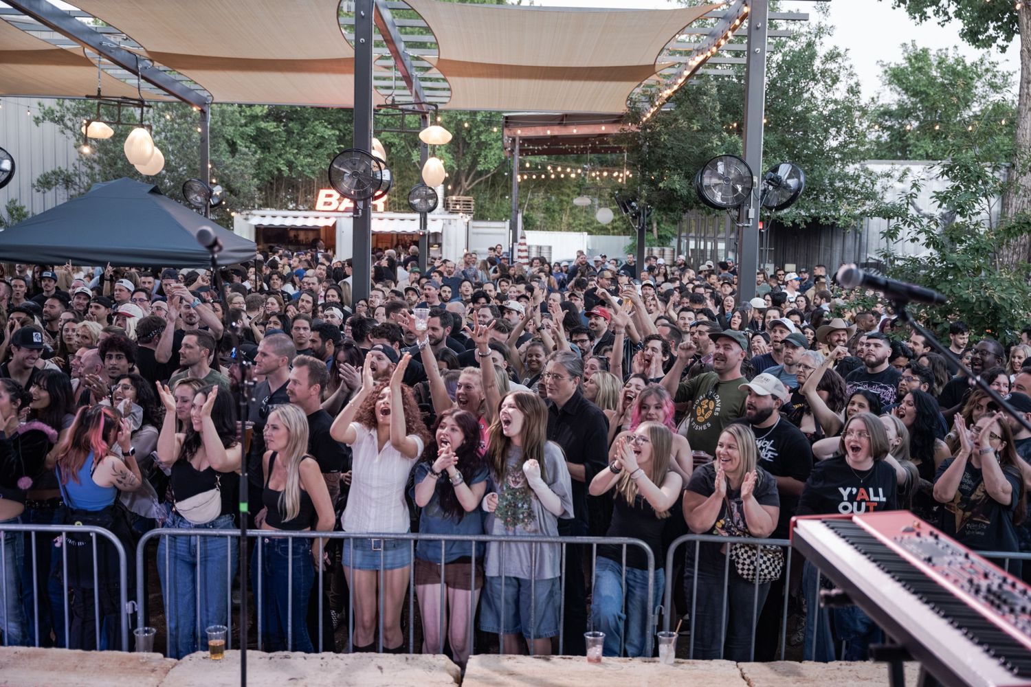 Large crowd of fans clapping at an outdoor concert venue with barriers and trees in background.