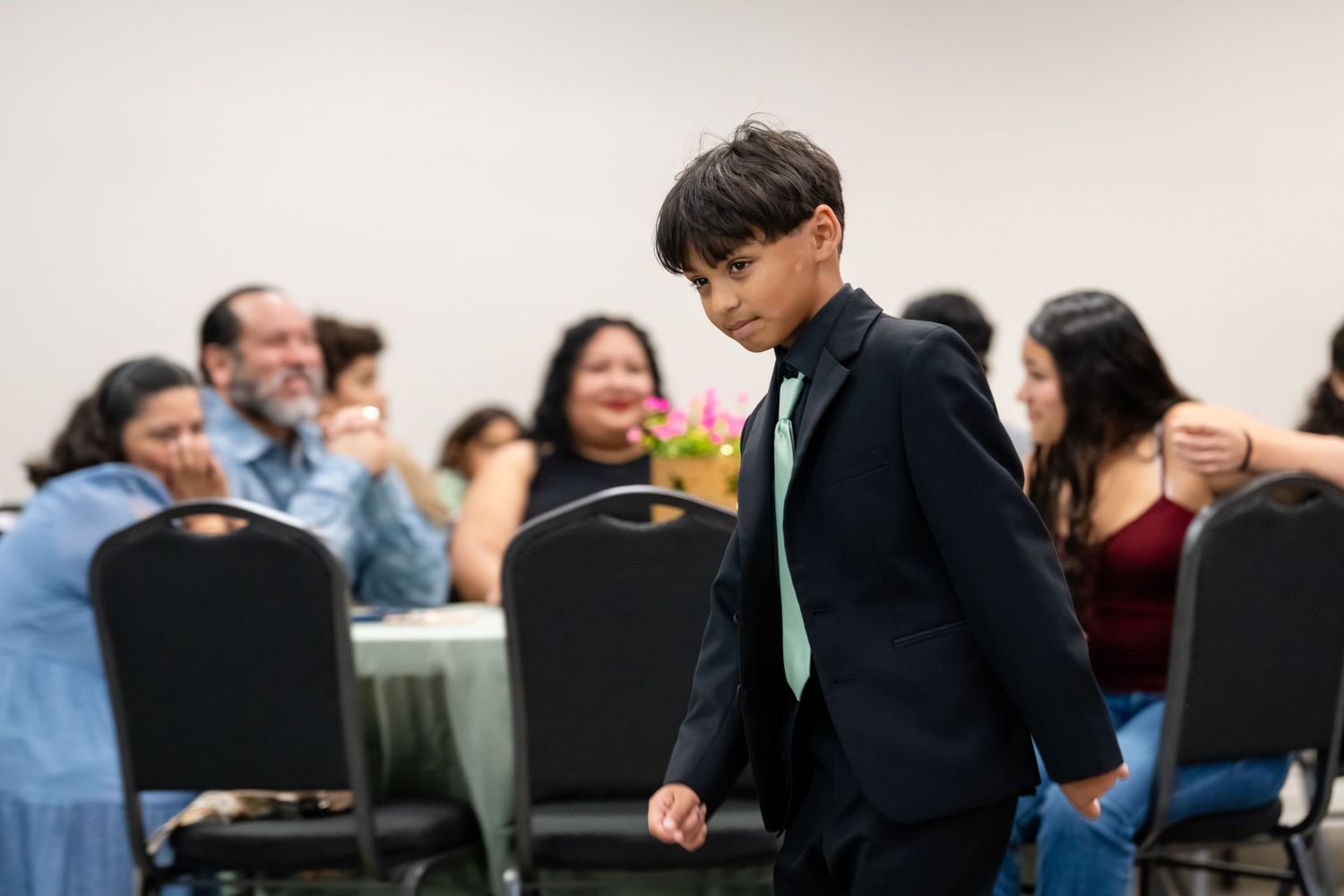 Young boy in black suit with green tie walks at formal banquet event with guests seated at tables.
