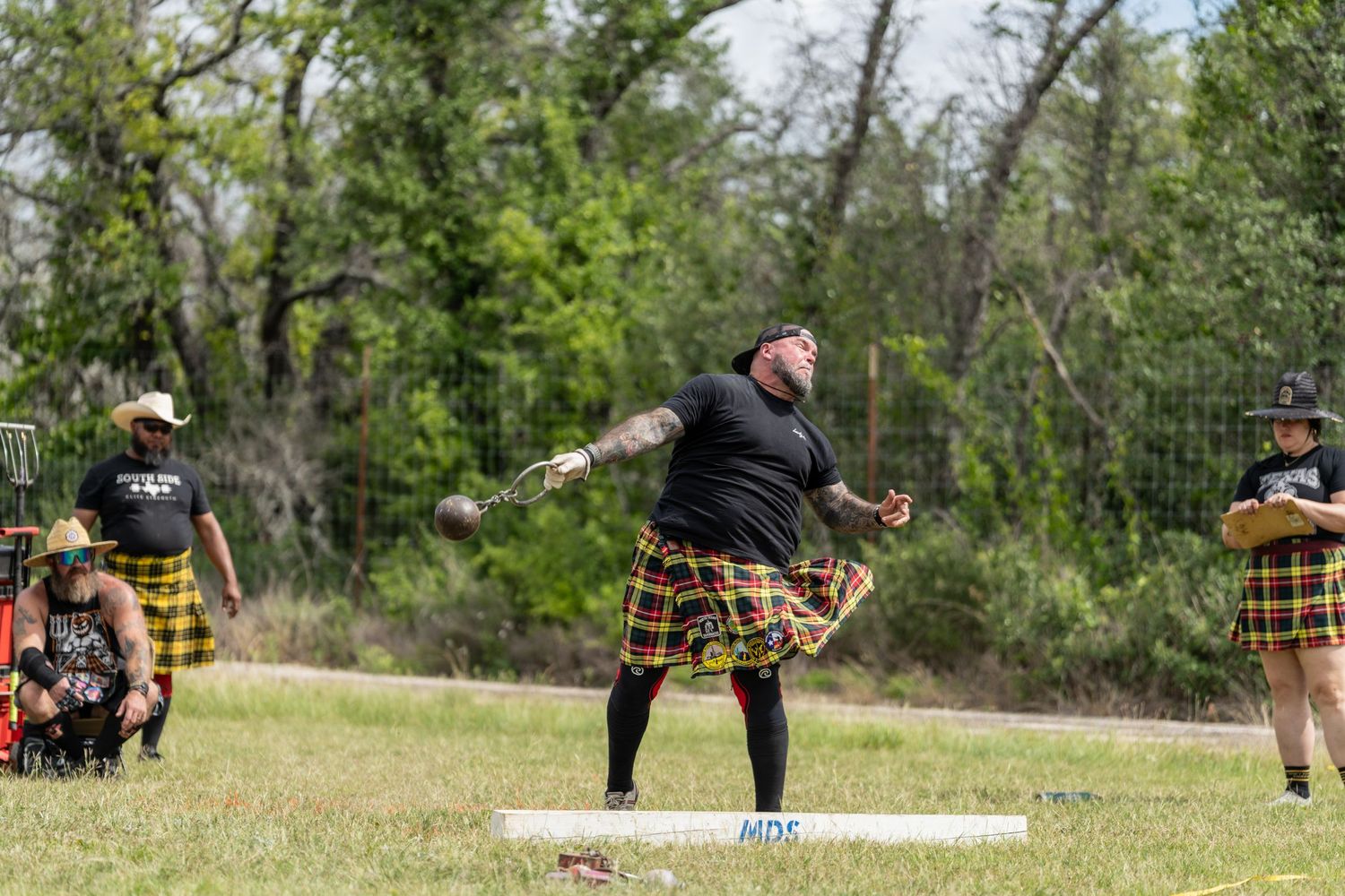 Man in tartan kilt competing in Highland Games hammer throw event outdoors with trees in background.