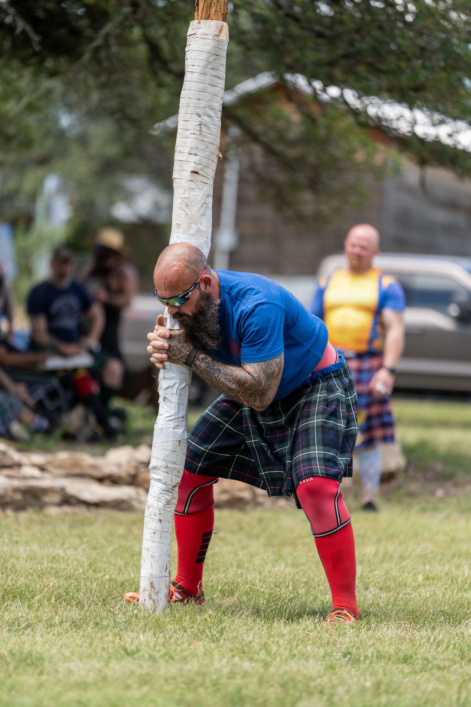 Tattooed man in blue shirt, tartan kilt, and red socks competing in caber toss at Highland Games.