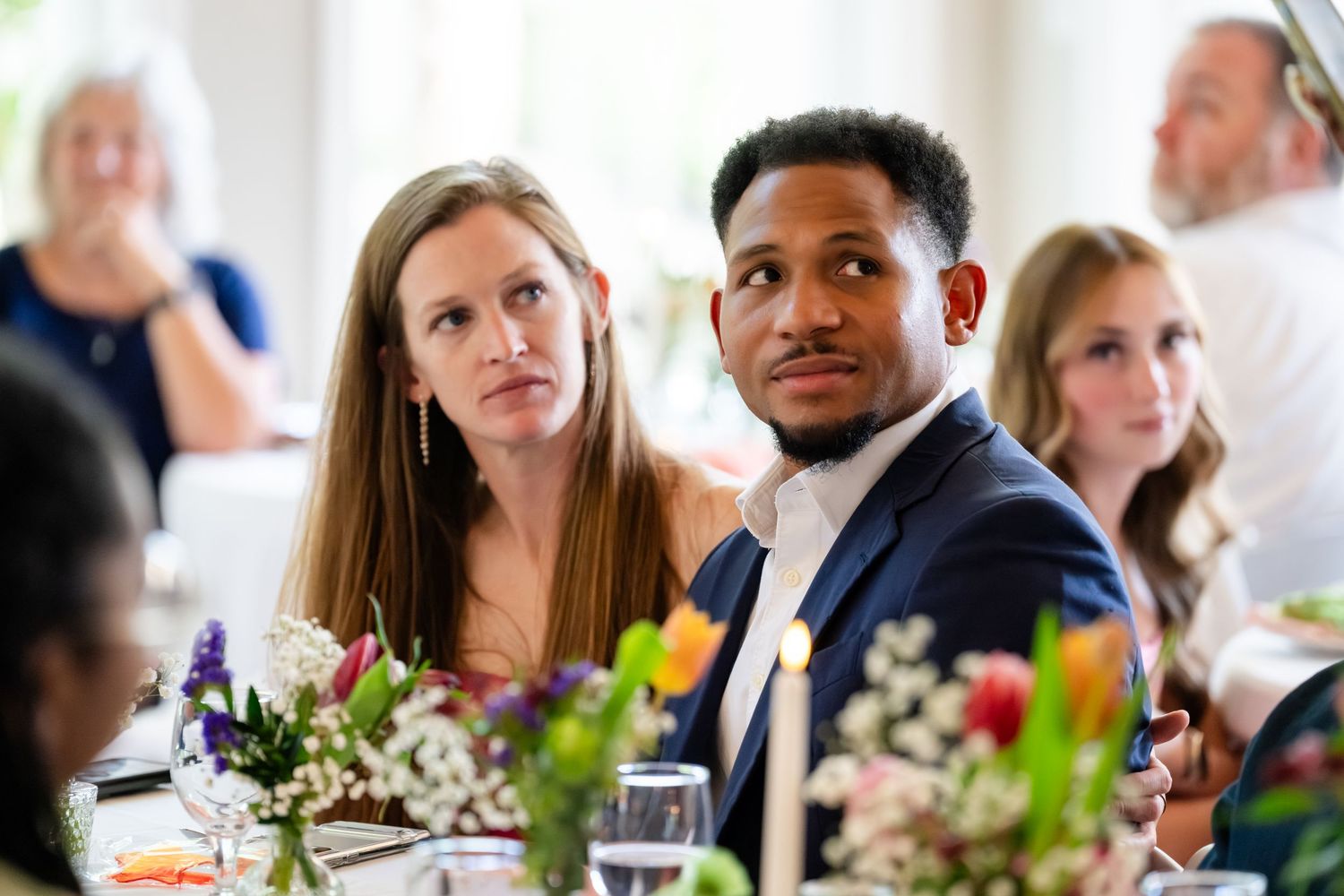 Couple seated at a floral-decorated wedding reception table, listening attentively during the event.