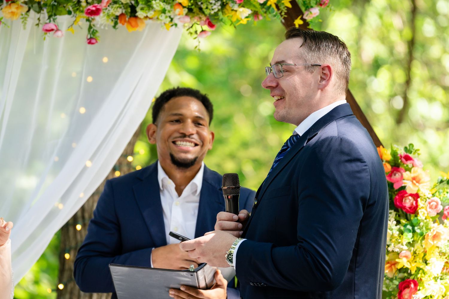Two grooms share a joyful moment during an outdoor wedding ceremony adorned with colorful floral decorations.