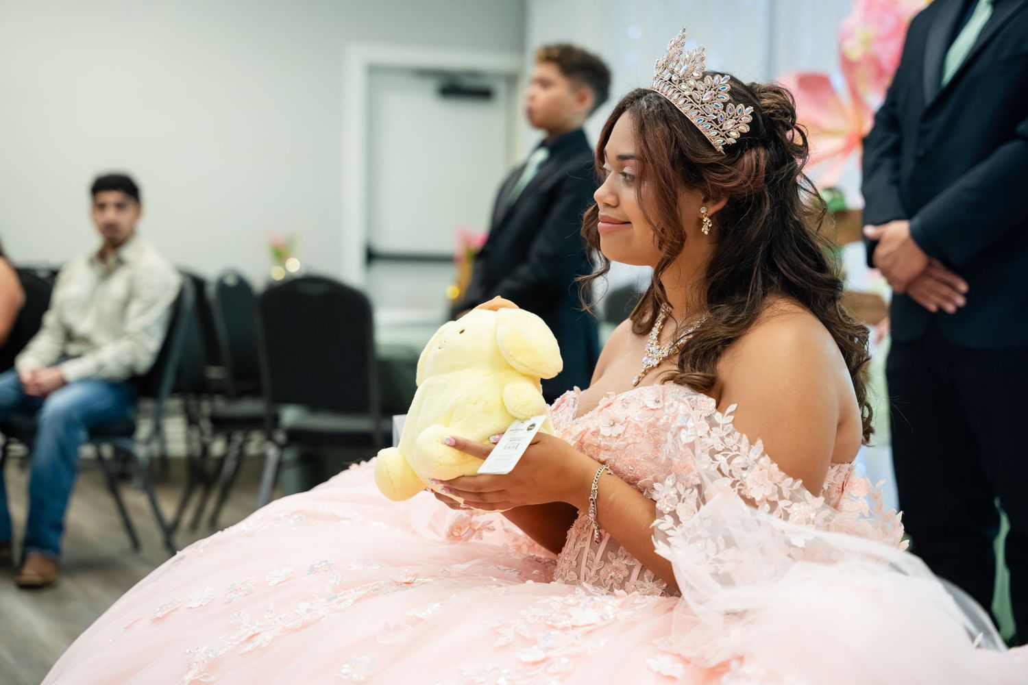 Young woman in pink quinceañera gown wearing tiara holds yellow stuffed animal at her celebration party.