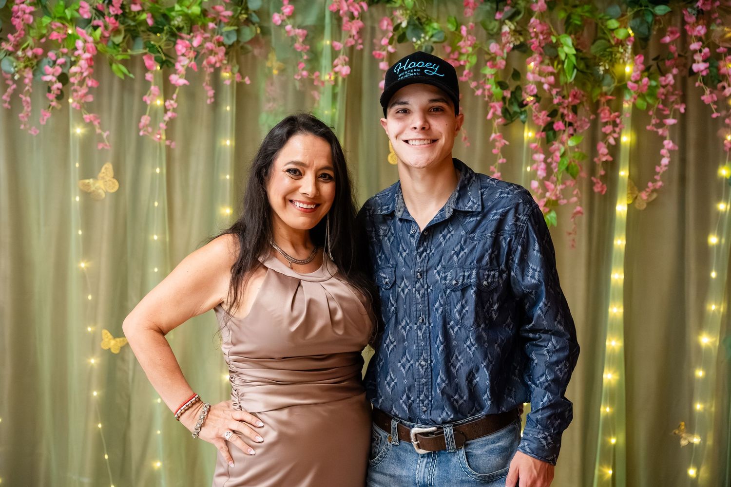 Couple posing together in front of floral backdrop with pink flowers and string lights at a formal event.