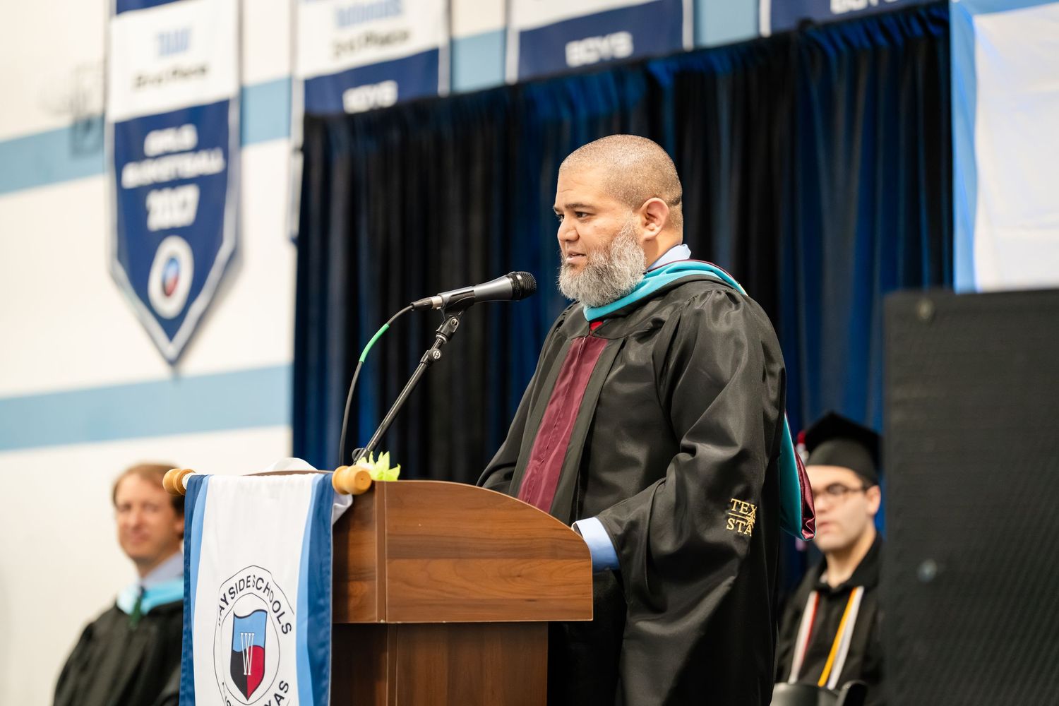 Man in graduation regalia speaks at podium during academic commencement ceremony in school gymnasium.