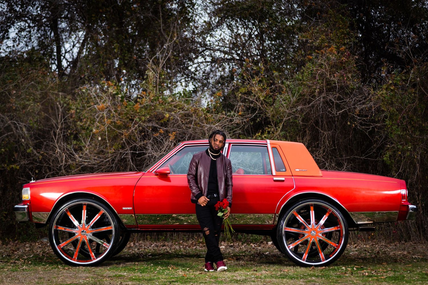 A red classic car with oversized custom wheels sits parked on a dirt road surrounded by autumn trees.