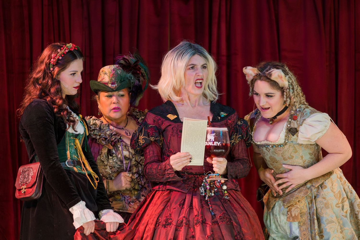 Theatrical performers in elaborate period costumes act on stage with red curtains in the background.