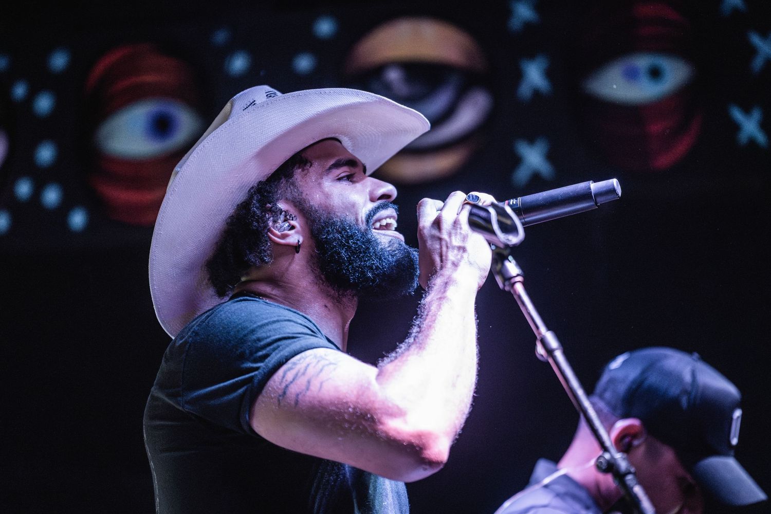 A performer in a white cowboy hat sings into a microphone on stage with blue lighting and decorative backdrop.