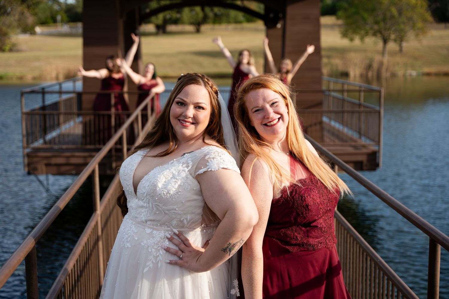 Two people in wedding attire pose playfully on a wooden bridge over a lake during sunset.