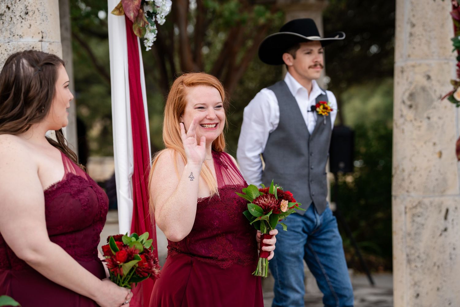Wedding party members in burgundy dresses and western attire gather near floral-decorated stone pillars.