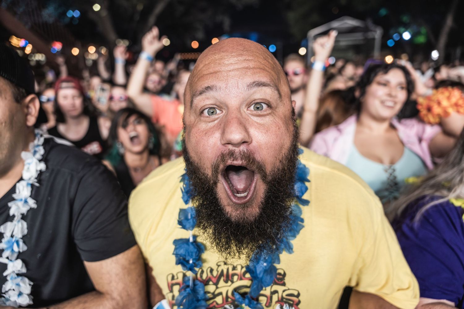 An excited festival attendee in a yellow shirt with a blue lei reacts enthusiastically in a crowd of concert-goers.