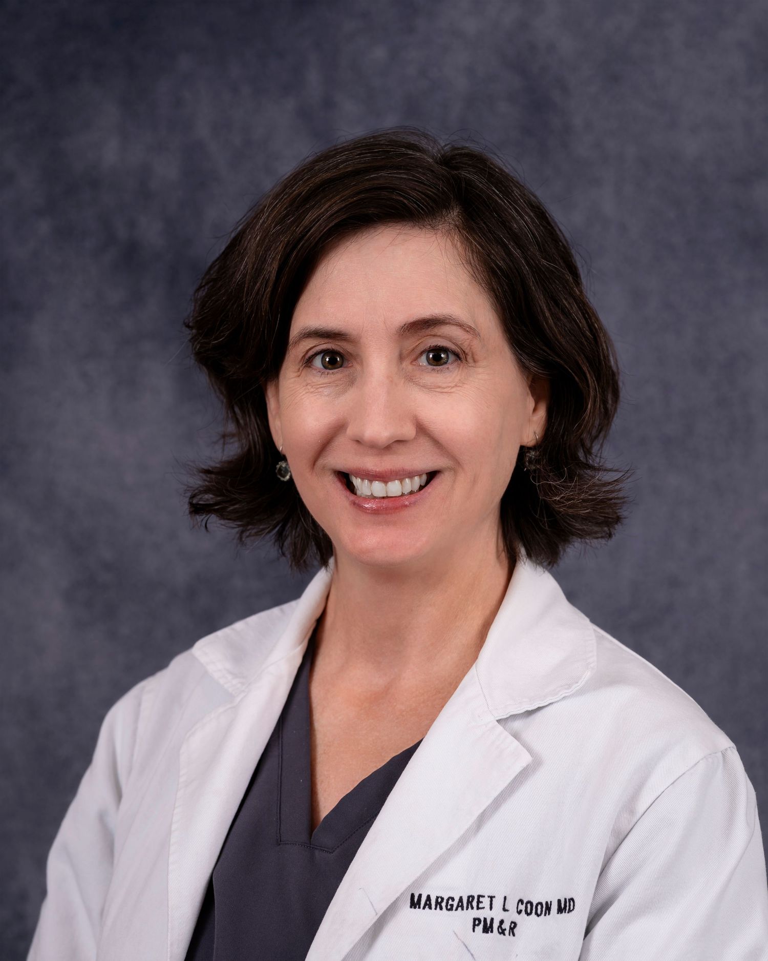 Professional medical office portrait of a doctor wearing a white coat and gray scrubs against a dark background.