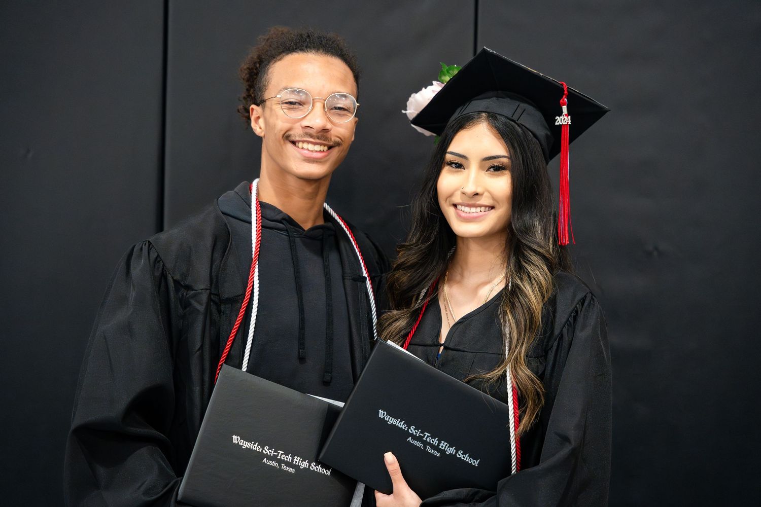 Two graduates holding diplomas pose together against a black backdrop in traditional black caps and gowns.