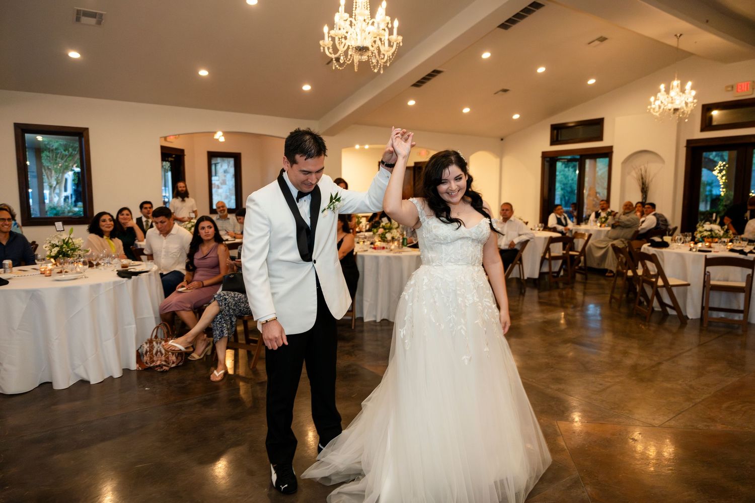 Dancers glide across a warm-lit banquet hall floor during an elegant wedding reception as guests watch from dining tables.