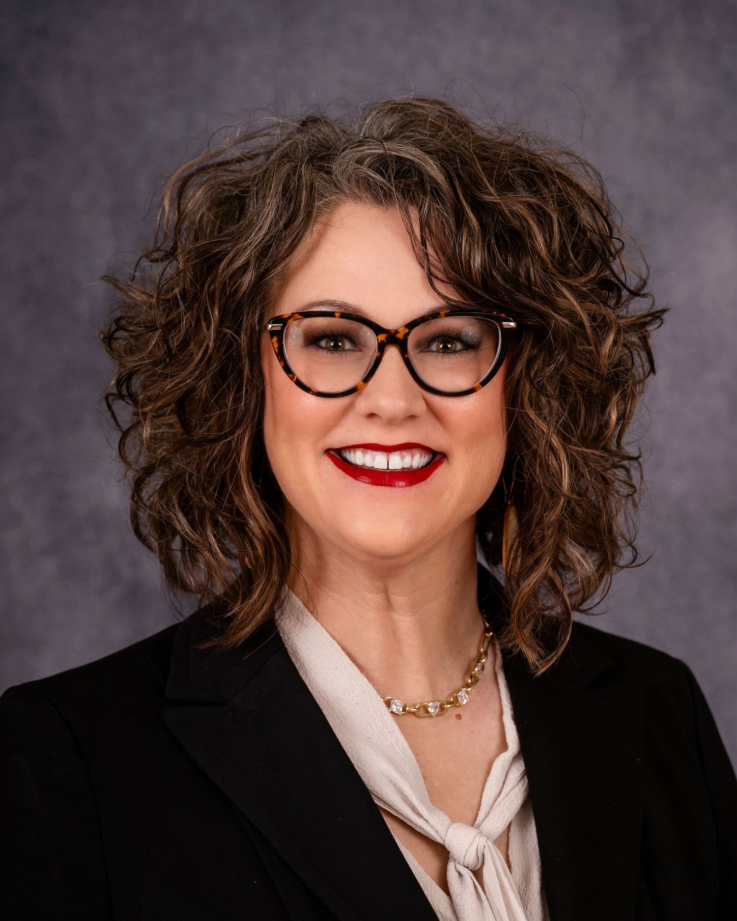 Professional headshot featuring curly brown hair, cat-eye glasses, red lipstick, and a black blazer with white blouse.