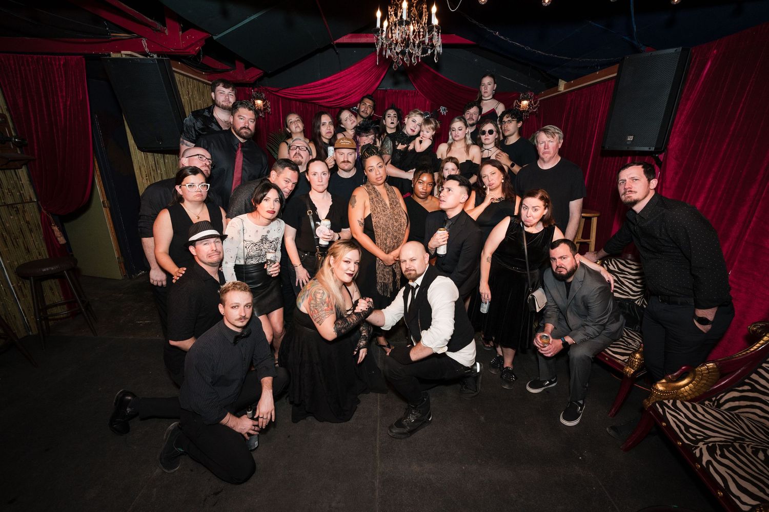 Large group photo of party guests in black attire posing in a dimly lit venue with red decor.