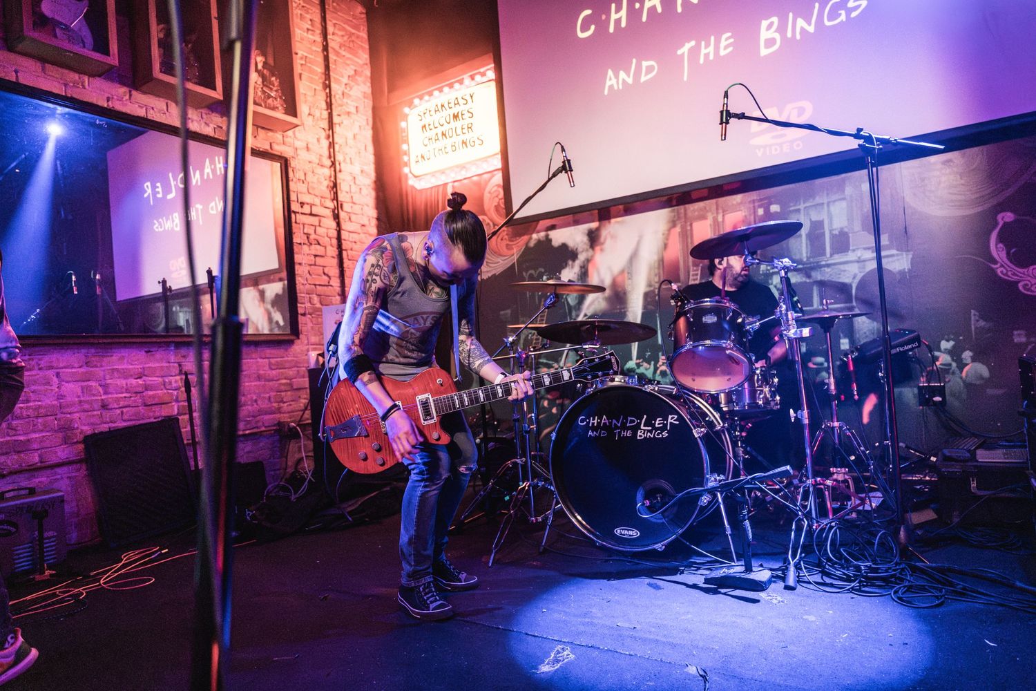 A wide shot of a concert stage with drums and instruments set up under blue and red lighting.