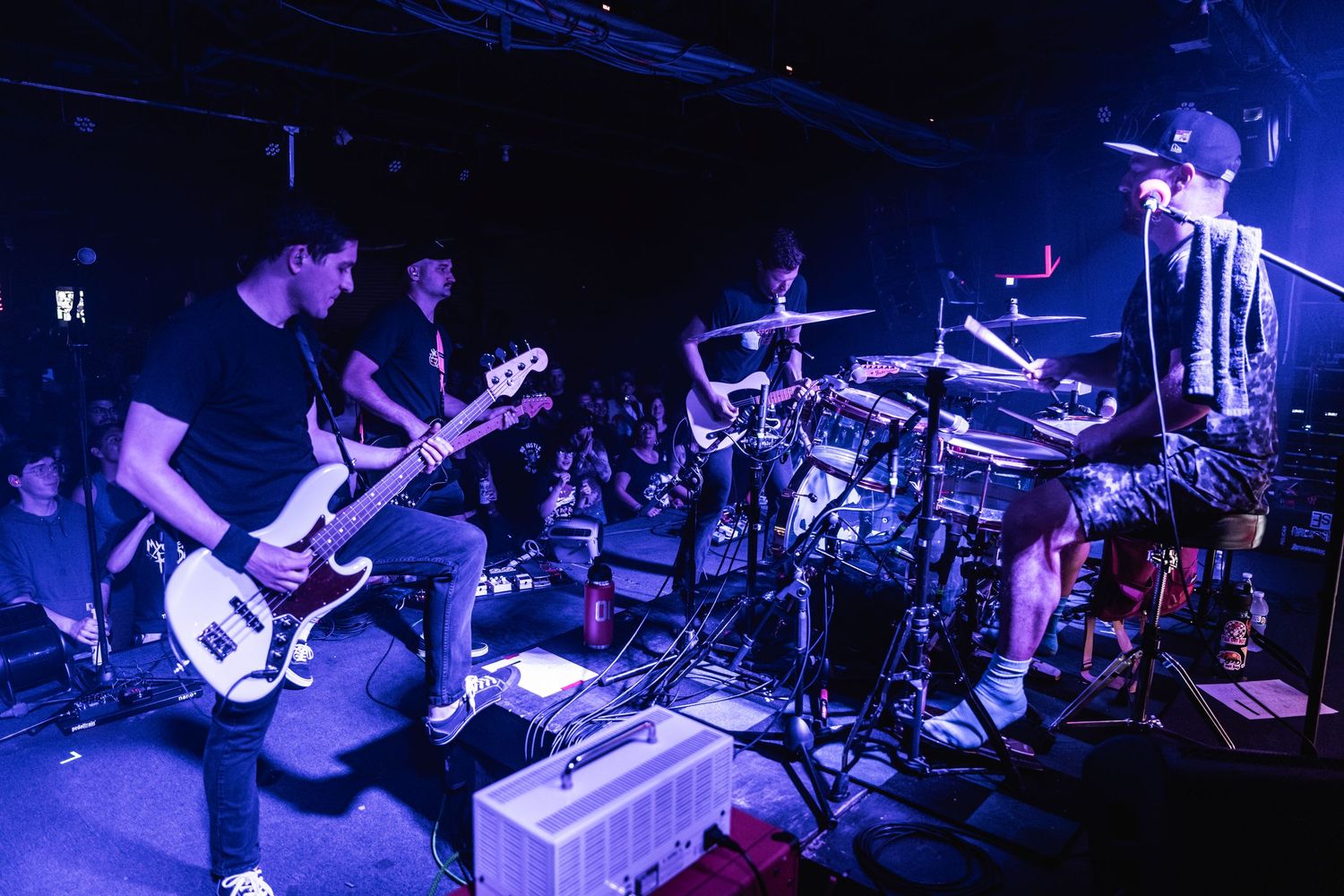 A band performs on stage at a dark venue with blue lighting while enthusiastic concert-goers watch from the crowd.