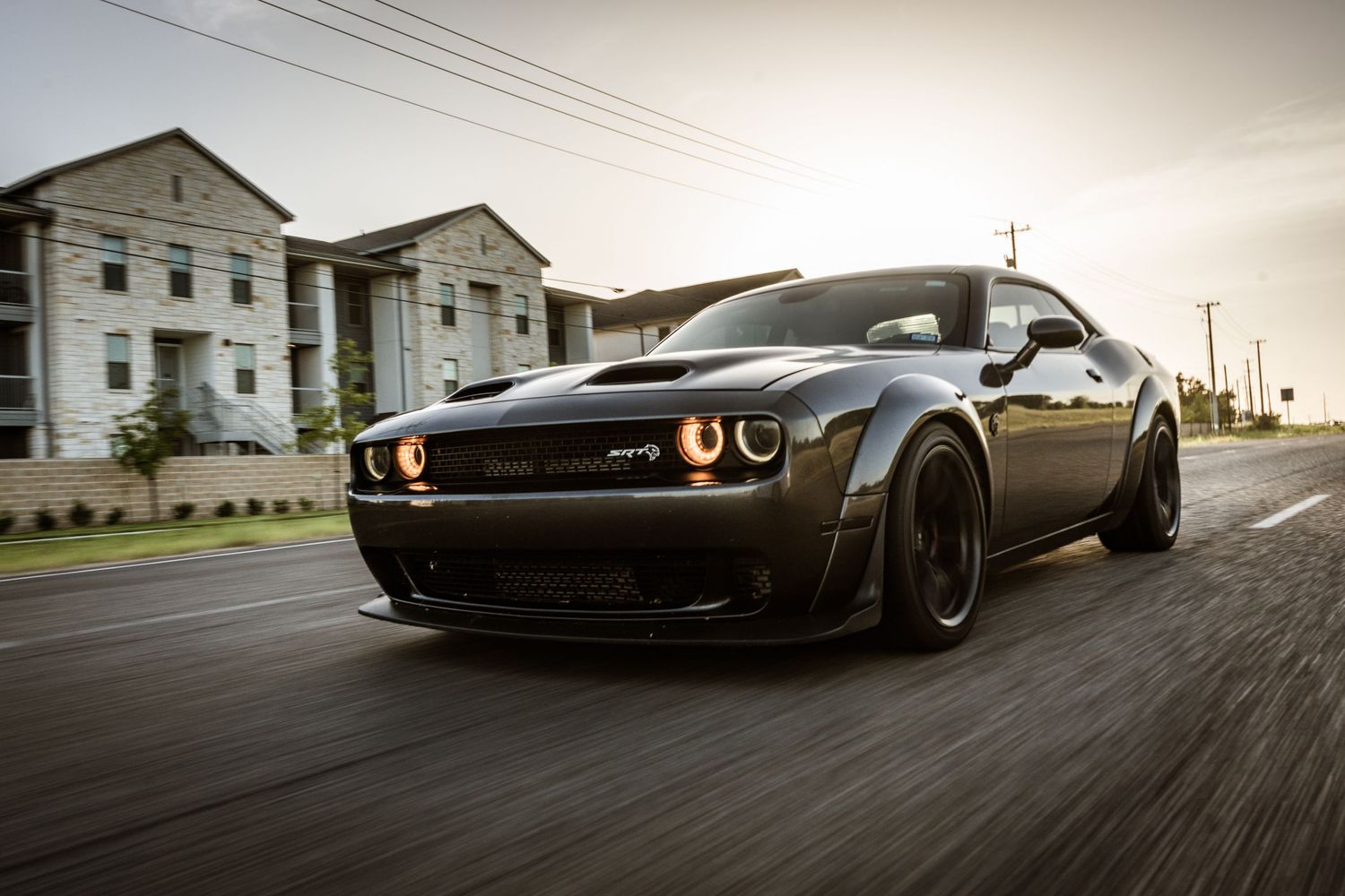 Black Dodge Challenger SRT Hellcat with glowing orange halo headlights racing on a street at dusk.