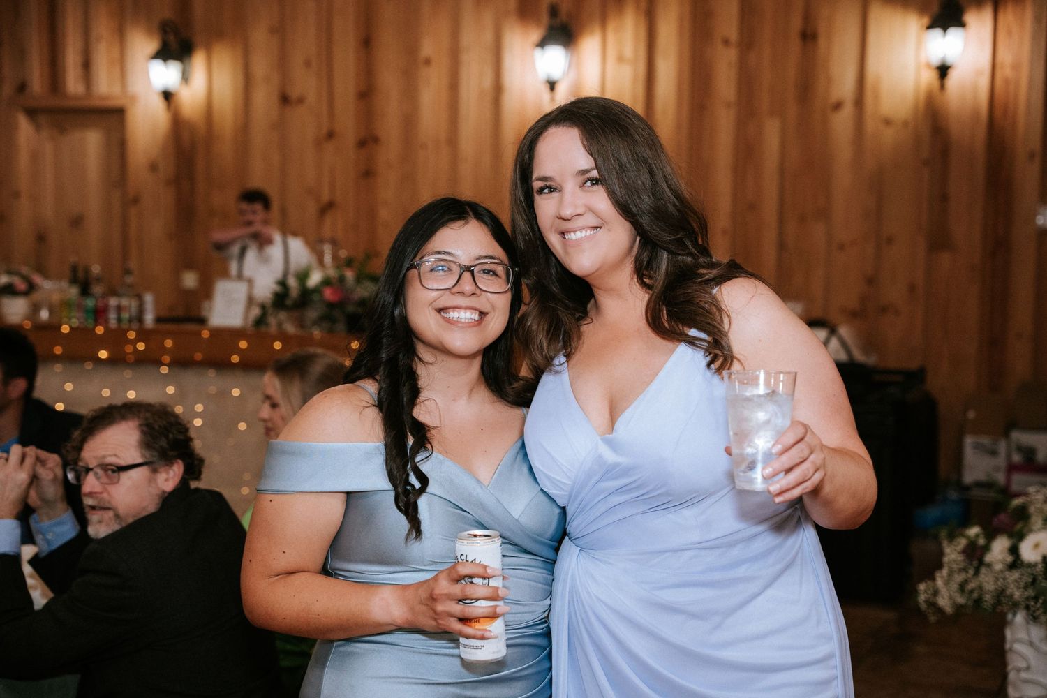Two friends in blue dresses share a laugh while holding drinks at a rustic wedding reception.