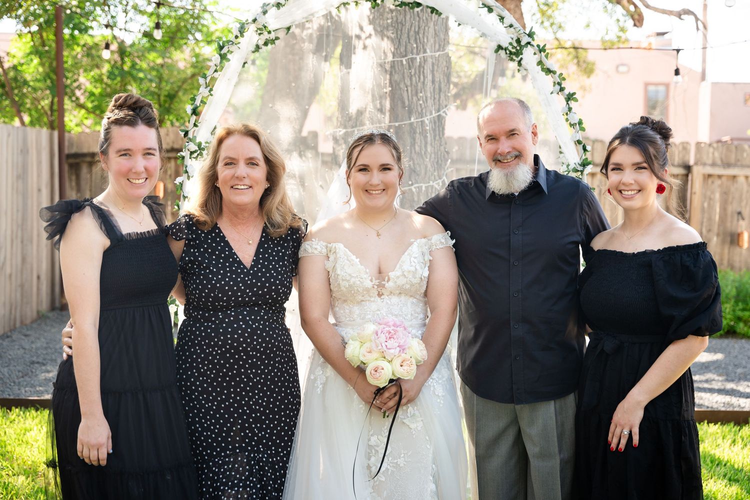 Wedding party members pose together in front of a wooden fence and floral arch on a sunny day.