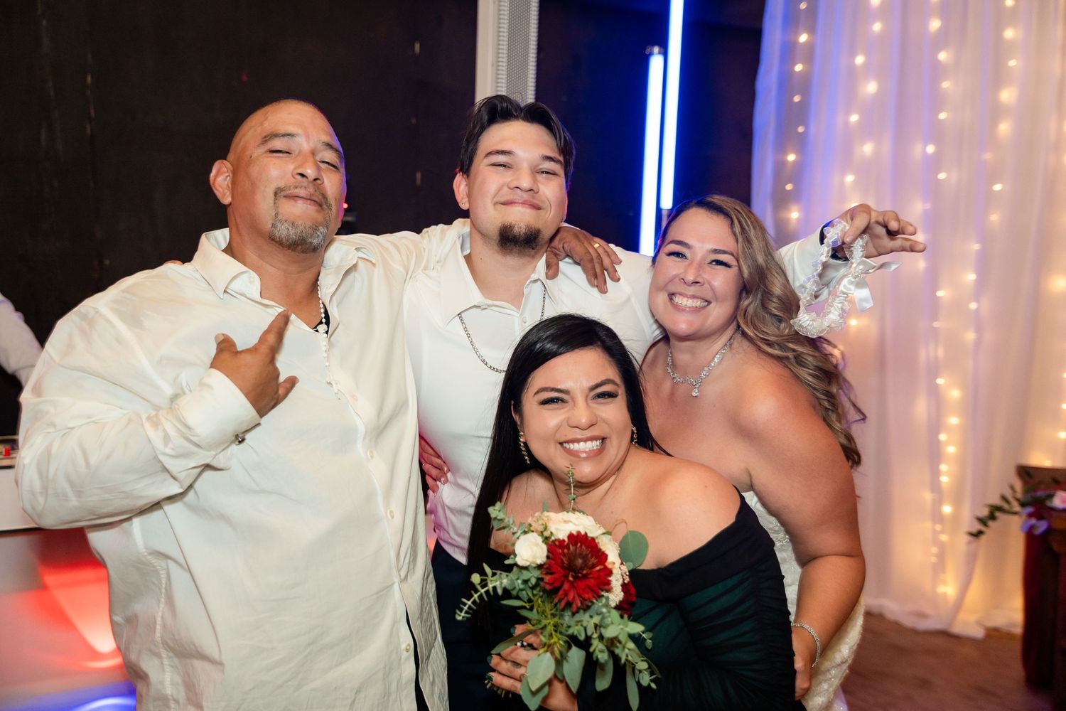 A group photo of wedding guests posing together with bouquets under string lights.
