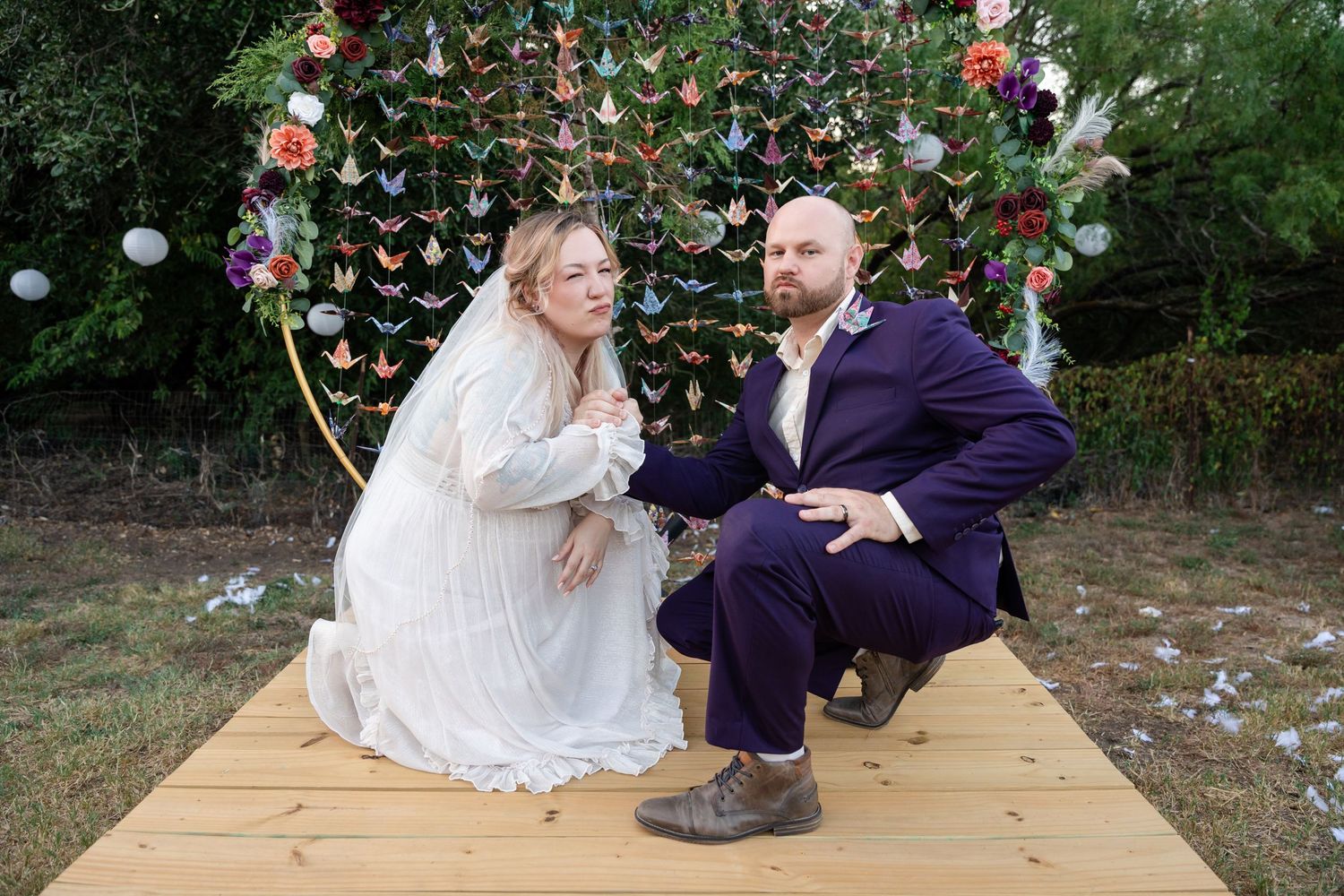 A couple kneels on a wooden platform under an arch decorated with paper butterflies and flowers.