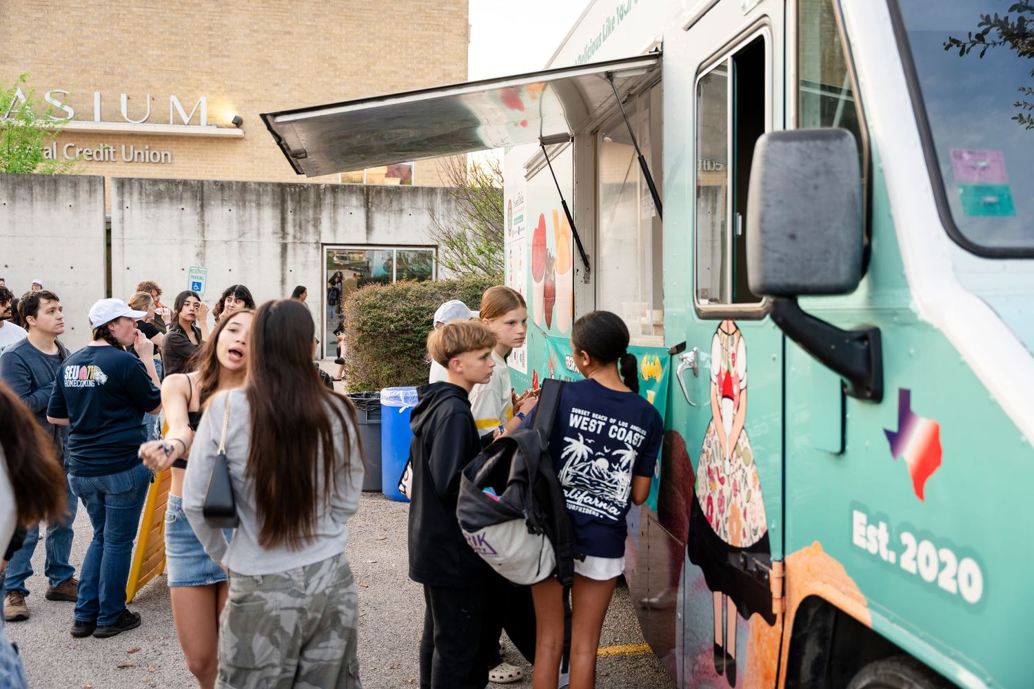 A line of customers waiting to order from a food truck parked outside on a sunny day.